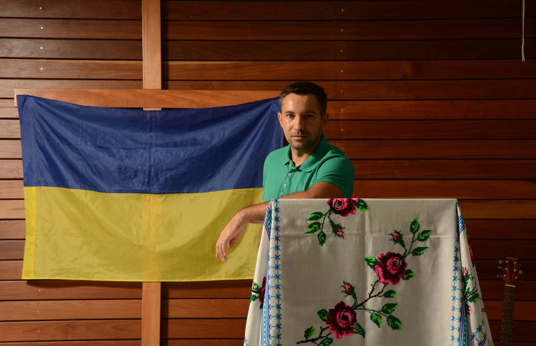 Homem de camisa verde, apoiado a púlpito e em frente a uma bandeira da Ucrânia pendurada na parede de madeira