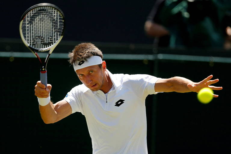 Homem branco de cabelo escuro, curto e liso, de roupas brancas acerta uma bolinha de tênis com uma raquete. 