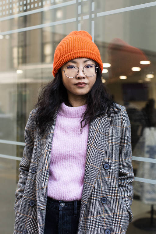 mulher com traços asiáticos, sobretudo cinza, touca laranja e uma blusa rosa em frente a prédio espelhado