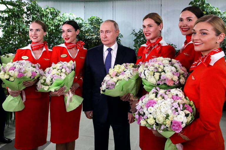 O presidente russo, de terno e gravatas escuras e camisa branca, possa em pé ladeado por duas comissárias do lado direito dele e três do lado esquerdo, todas de vermelho vivo e segurando um buquê de flores brancas e roxas