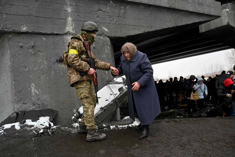 Soldado ucraniano ajuda idosa a atravessar uma ponte destruída enquanto ela deixa a cidade de Irpin, a noroeste de Kiev