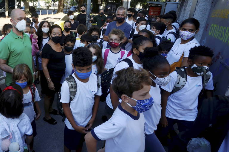 Crianças de camiseta branca estão agrupadas na calçada, de máscara, fazendo uma curva em direção ao que parece ser o portão da escola. Alguns adultos, também de máscara, observam