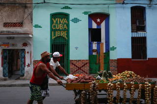 Two men pull a cart with fruits and vegetables for sale as they pass by the Cuban flag and the phrase 'Viva Cuba Libre' (Long Live Free Cuba), painted on the doors of a building, on a street in Havana
