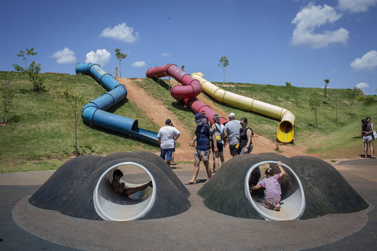 Três tobogãs, um azul, um vermelho e um amarelo, estão dispostos em um pequeno relevo; abaixo, há dois túneis, onde brincam crianças