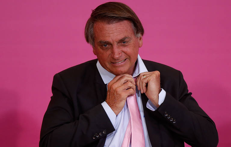 À frente de um fundo rosa, Jair Bolsonaro (PL), de frente e de terno preto, desafoga nó de uma gravata rosa; foto foi feita em cerimônia no Palácio do Planalto em homenagem ao Dia Internacional das Mulheres, em março deste ano