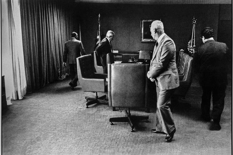 quatro homens de pé, prestes a se sentar, em sala de escritório