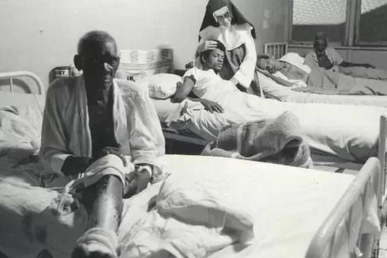 Irmã Dulce abraça um homem deitad em um maca de hospital. Ao redor há outras macas e pacientes.