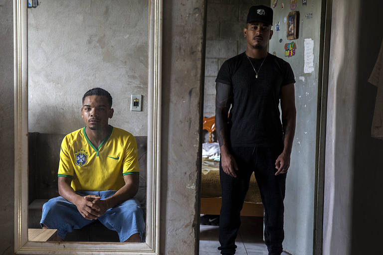 No canto esquerda, se vê o reflexo em um espelho de um garoto negro usando a camiseta da seleção brasileira. À direita, em pé em uma porta, está outro garoto negro de roupa preta e boné. Ambos estão sérios.