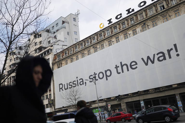Homem agasalhado passa por um prédio com uma faixa de protesto que diz em inglês: "Rússia, pare a guerra!"