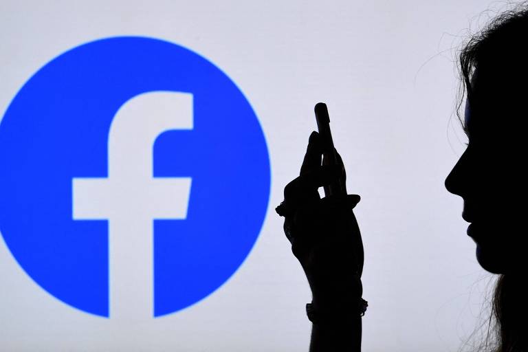 Foto ilustrativa mostra silhueta de pessoa cim smartphone na mão, à frente de logo do Facebook em parede branca