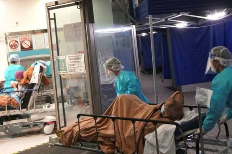 Dois profissionais da saúde empurram uma maca, com um paciente deitado nela, para dentro de uma sala