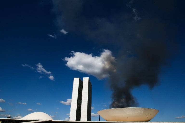 Dois prédios altos parelos, uma cúpula voltada para baixo e outra voltada para cima (os prédios do congresso nacional) tendo ao fundo um céu azul e uma coluna de fumaça