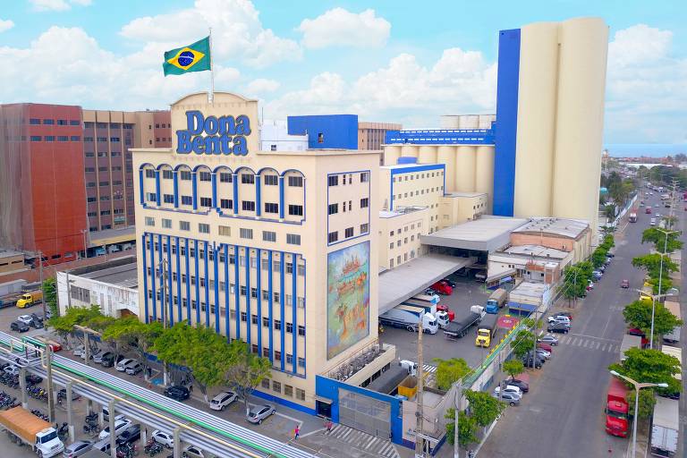 prédio de cinco andares na cor bege, com detalhes em azul, com o logotipo D. Benta e uma bandeira do Brasil no topo
