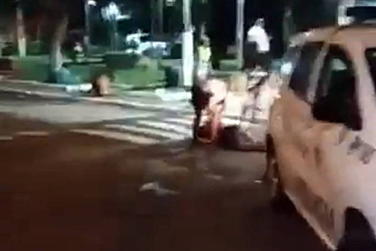 reprodução de vídeo mostra detalhe de viatura da polícia branca com corpo de homem no chão ao fundo