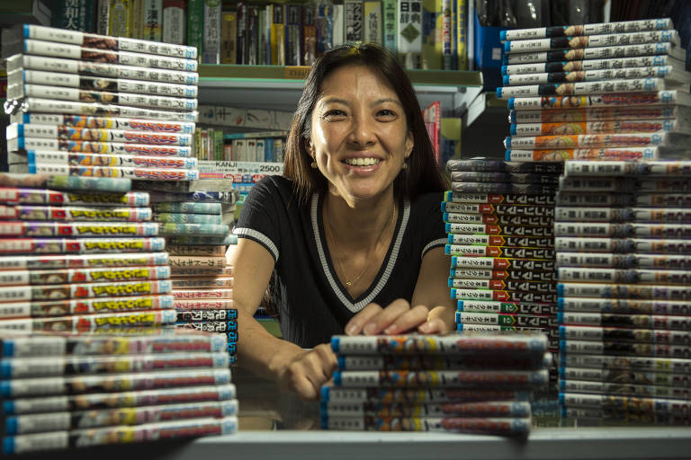 Imagem destaca pilhas de livros em japonês sobre uma mesa, ao redor de Tatiana, que apoia as mãos sobre alguns livros; ela é uma mulher com cabelos lisos castanhos e olhos puxados, veste uma camiseta preta e sorri