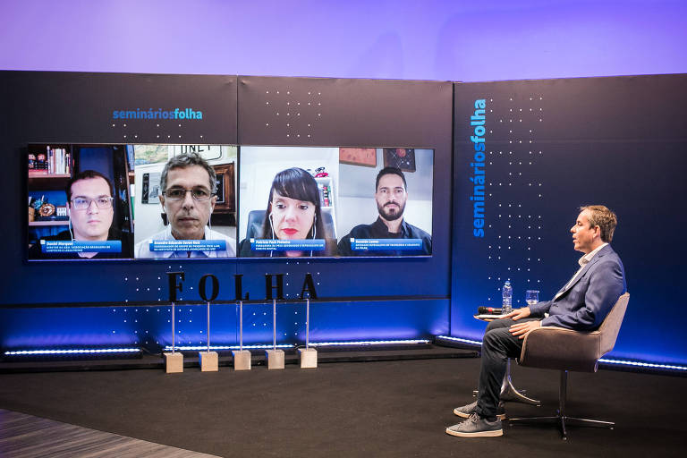Imagem mostra evento ocorrendo em um auditório; há um painel azul, no qual estão as imagens de quatro convidados que participam online; à direita, o mediador está sentado em uma cadeira