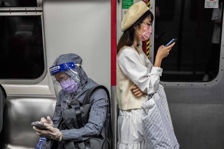 Pessoas de máscara em um trem. Uma está sentada e a outra, de pé.