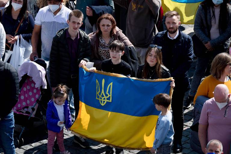 Da esquerda para a direita, uma menina de cerca de dois anos, um adolescente, uma mulher, um menino e uma menina de cerca de 12 anos e um menino de cerca de quatro anos carregam a bandeira da Ucrânia, azul e amarela, no centro de uma praça. 