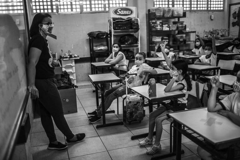 Professora usa máscara em sala de aula, à esquerda da imagem encostada na lousa; alunos ocupam carteiras da sala e alguns levantam a mão durante exercício