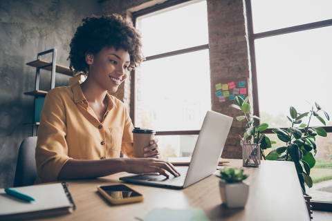 Profile side photo of charming smart afro american girl entrepreneur use computer, work training company career development seminar sit table hold takeout mug hot beverage in loft office. Credit Deagreez / Adobe Stock