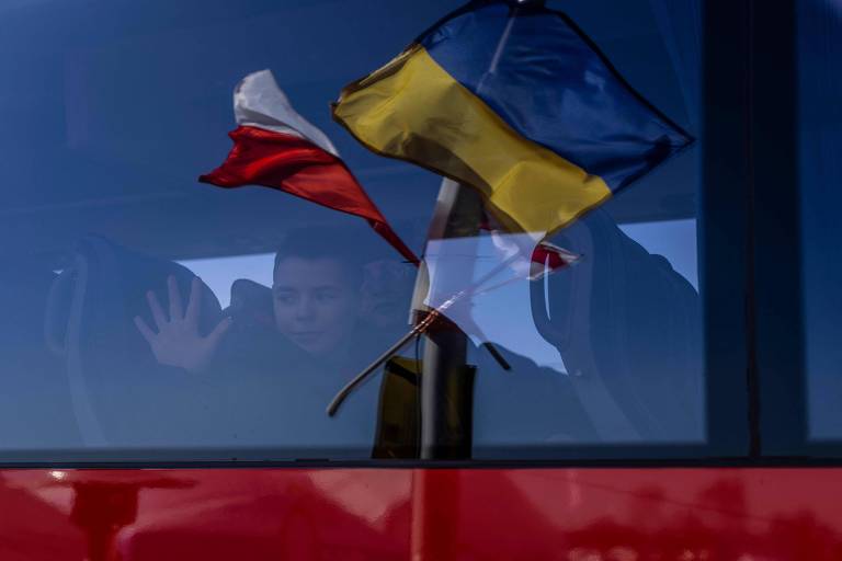 As bandeiras polonesa e ucraniana refletidas na janela de um ônibus, do qual um menino acena depois de chegar à Medika, na Polônia, fugindo da guerra na Ucrânia