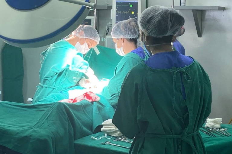 sala de cirurgia com paciente com roupas verdes na maca cercado de três profissionais de saúde com avental e toucas