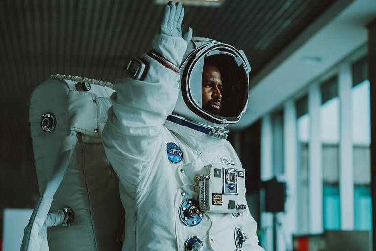 homem negro vestido de astronauta levanta mão em saguão de aeroporto