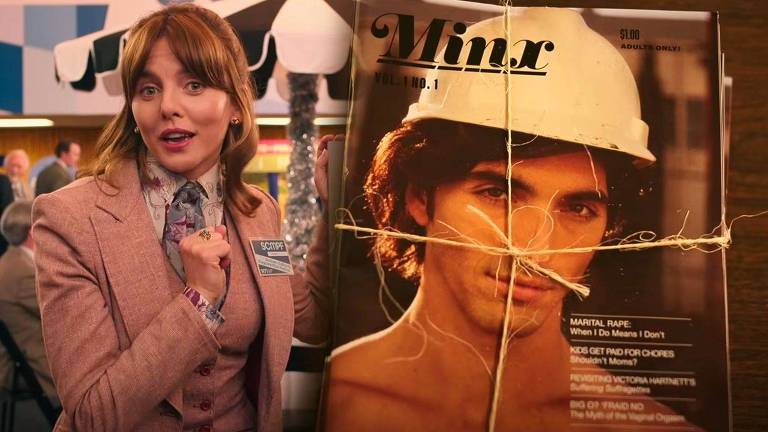 Cena de seriado que retrata uma mulher vestida com um blazer bege ao lado de uma grande capa de da revista fictícia Minx, que estampa um homem bonito com um chapéu de constrção