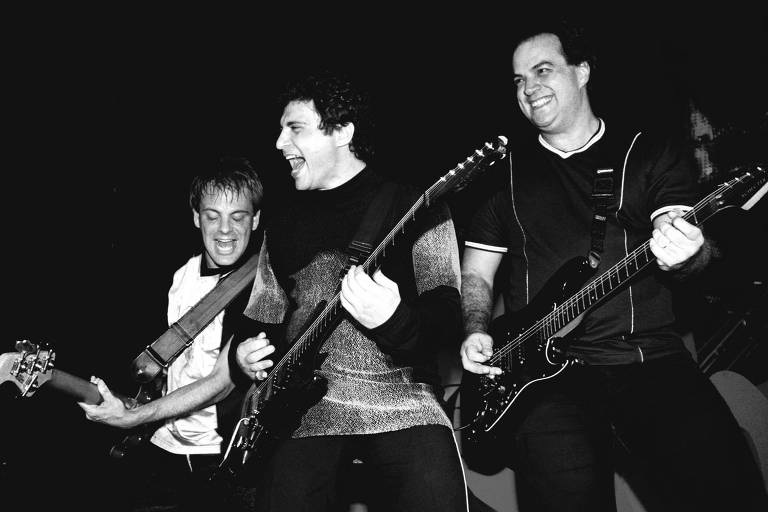 Foto em preto e branco. Trio de homens brancos com guitarra em palco. 