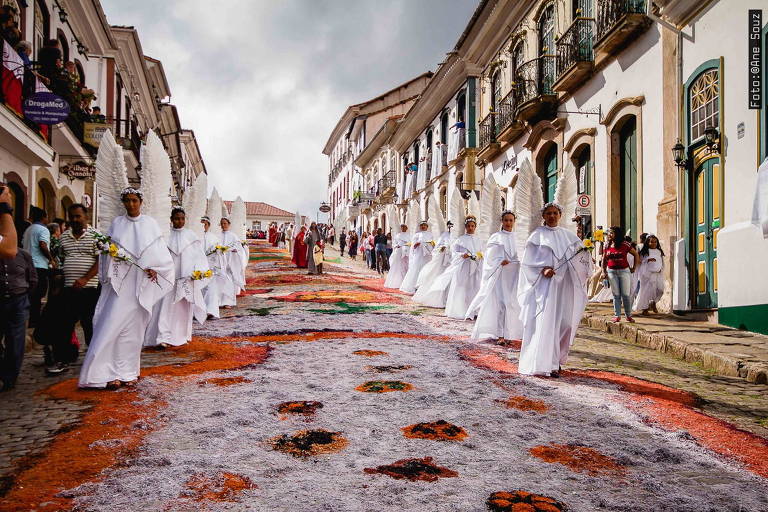 Tapetes coloridos enfeitam a rua, enquanto pessoas com roupas de anjo participam da procissão