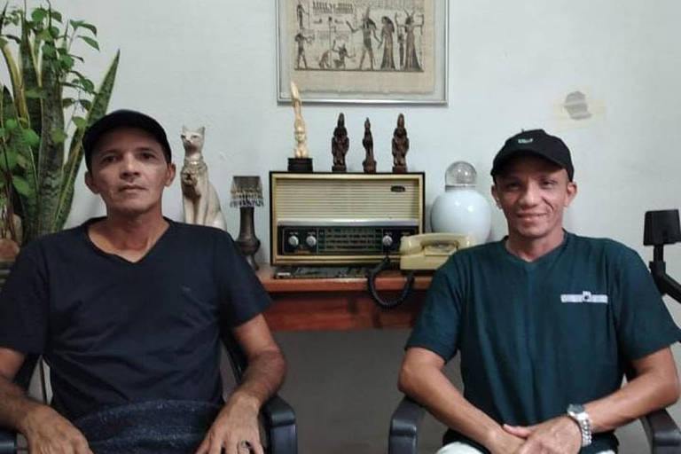 Dois homens sentados, de boné e camiseta pretas. Atrás, quadro egípcio e esculturas do Egito em cima de um rádio antigo em um aparador