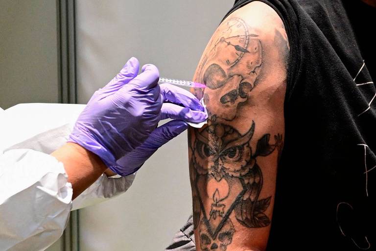 Um jovem recebe a vacina em um centro de vacinação em Berlim. Só aparece seu braço direito, além das mãos do profissional de saúde que injeta o imunizante. Ele tem uma tatuagem de coruja em traços pretos densos, e o profissional usa luvas.