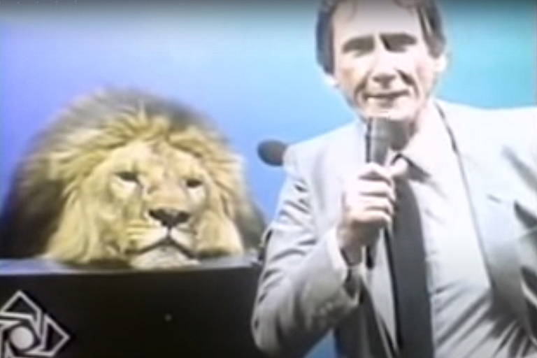 Homem com microfone na mão em estúdio, com um leão de verdade sentado em uma bancada atrás dele