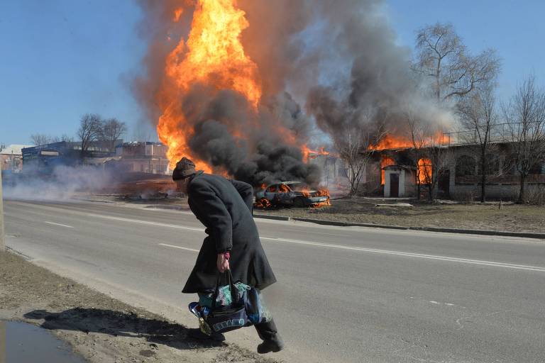 Homem com casado escuro e carregando uma sacola parece correr enquanto, ao fundo, uma casa e um carro estão em chamas.