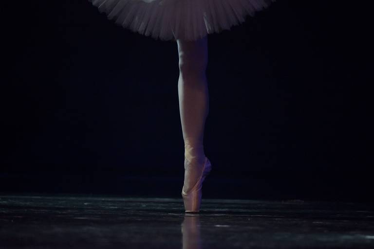 Perna de bailarina com sapatilha e tutu brancos com fundo preto.