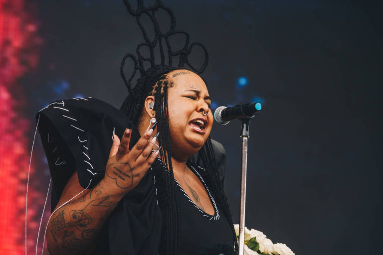 mulher negra trans com penteado excêntrico de tranças remetendo a uma coroa e onde se lê "Jup do Bairro" se apresenta em show