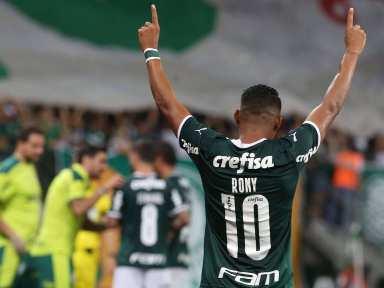 Jogador do Palmeiras (de costas) comemora gol no campo em meio a outros jogadores levantando os braços com os dedos indicativos em direção ao céu; equipe é vista ao fundo 