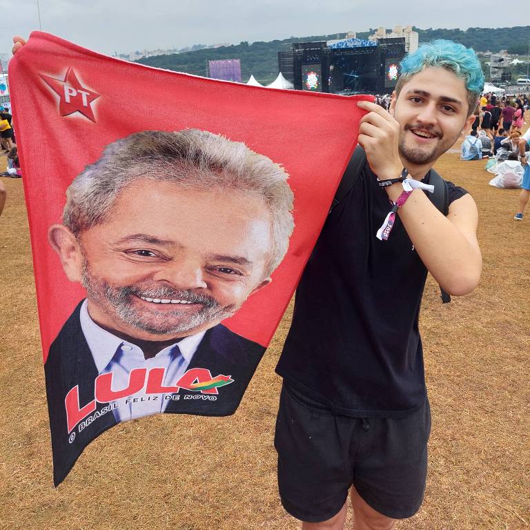Jovem de cabelos azuis exibe toalha vermelha com rosto do ex-presidente Lula (PT) em meio a um gramado