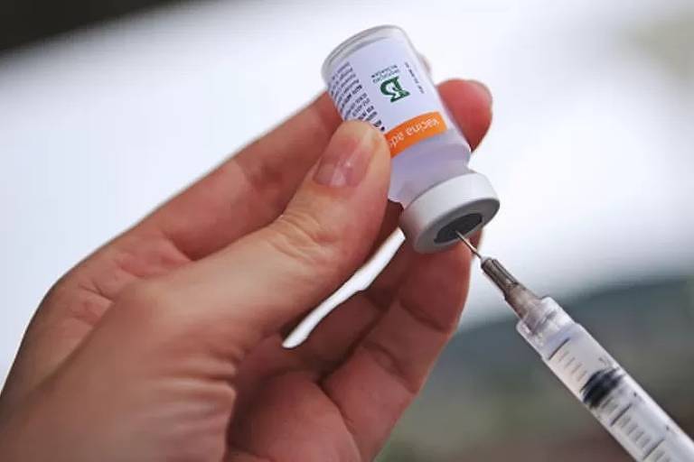 Imagem em close mostra as mãos de uma pessoa espetando uma seringa em uma ampola de vacina