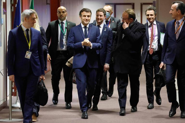 No centro, o presidente da França, Emmanuel Macron aperta as mãos enquanto caminha. Ele usa terno escuro e é acompanhado por sete homens, também de terno, que parecem ser de sua comitiva. 