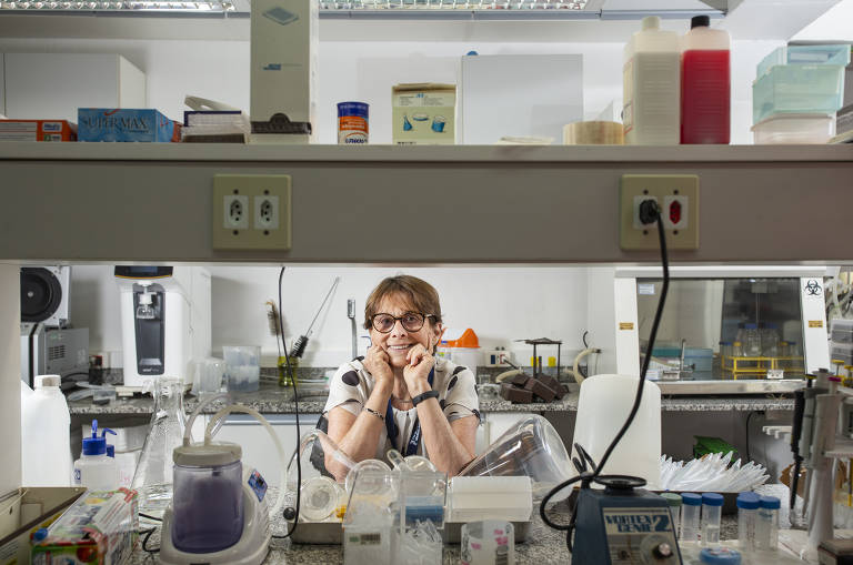 Mulher com as mãos debaixo do queixo posa em bancada de laboratório com diversas máquinas, equipamentos e reagentes ao seu redor