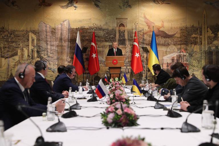 Imagem de uma grande mesa, com negociadores russos de um lado e ucranianos de outro; Recep Tayyip Erdogan está no fundo, no palco, e aparece ao centro. 