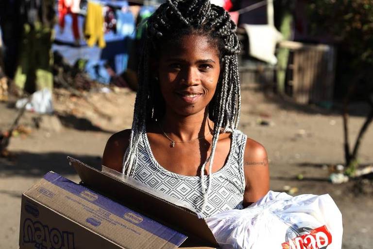 menina negra de cabelos trançados segura cestas básicas em rua arborizadas