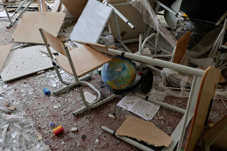 Imagem de uma sala de aula destruída, com cadeiras e mesas quebradas e espalhadas. No chão, um globo escolar aparece entre os destroços