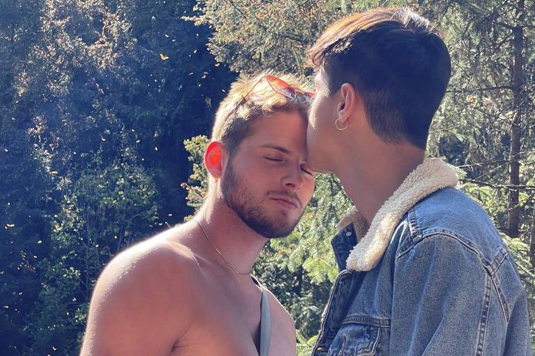 Dois homens brancos juntos, um beija a testa do outro, em frente a uma paisagem de topos de árvores