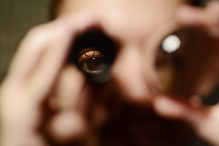 Um homem está com uma lupa nos olho esquerdo e uma lente redonda no olho direito. A imagem é fechada em seu rosto e é possível ver os olhos dele por meio dos objetos 