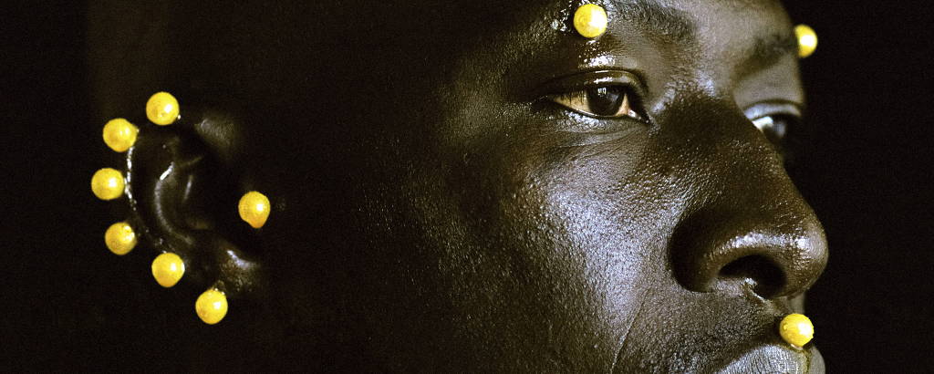 Fotografia do rosto de um homem negro, que tem esferas amarelas grudadas no lábio, no ouvido e nas sobrancelhas