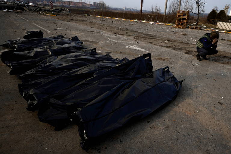 Cadáveres embalados em sacos pretos são dispostos em fila no chão; ao fundo, o agente de resgate de agacha