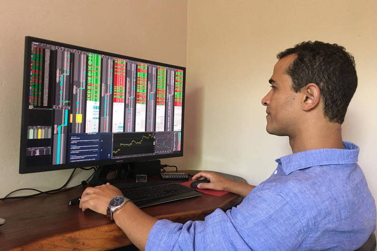 Moço de camisa azul sentado em frente a monitor com vários números de investimentos