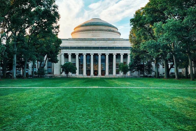 Foto mostra o prédio do MIT, uma construção clássica, em formato oval e branca, semelhante à Casa Branca. Na frente do instituto, há um extenso gramado, cercado por árvores.